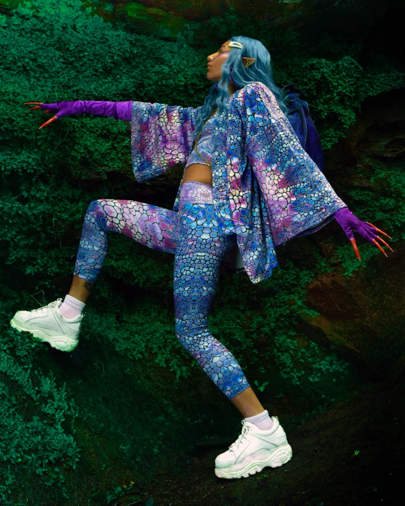 A model wearing a scaled purple and pink Kimono which is one of Kim's designs stands amongst mossy, clover-covered rocks. She has purple gloves with long orange claws, pixie-shaped ears, and blue hair. On her back, she has purple dragon wings.