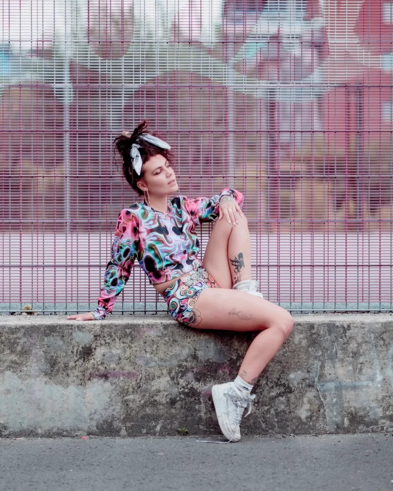A model wearing a cropped jumper and hotpants featuring Kim's textile prints looking off into the distance in an urban landscape. The mesh fence behind her has graffiti on it.