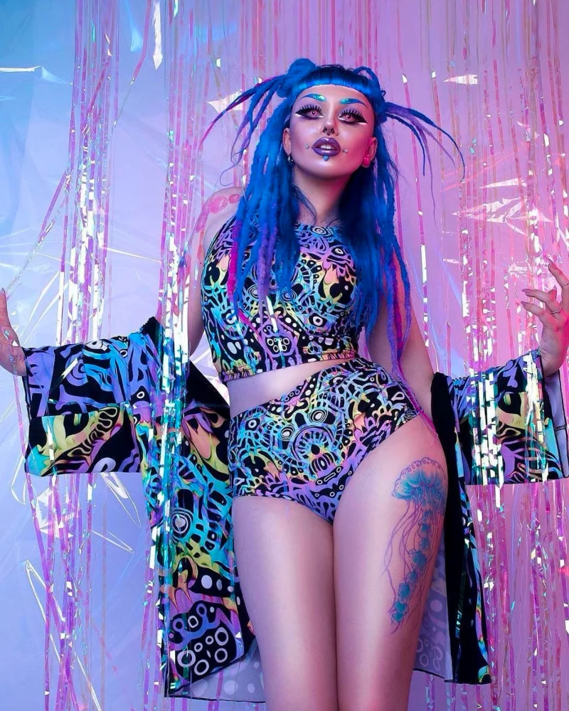 An alternative model wearing hotpants, crop top, and kimono featuring Kim's designs. The image has an underwater feel. The model is alternative with blue dreadlocked hair and facial piercings. On her thigh is a jellyfish tattoo.