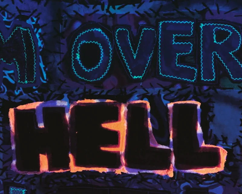 A close-up of the stitching details on Kim's artwork titled 'Prophecy' under ultra-violet light. The words say 'Over Hell' surrounded by glowing threads and fabric.