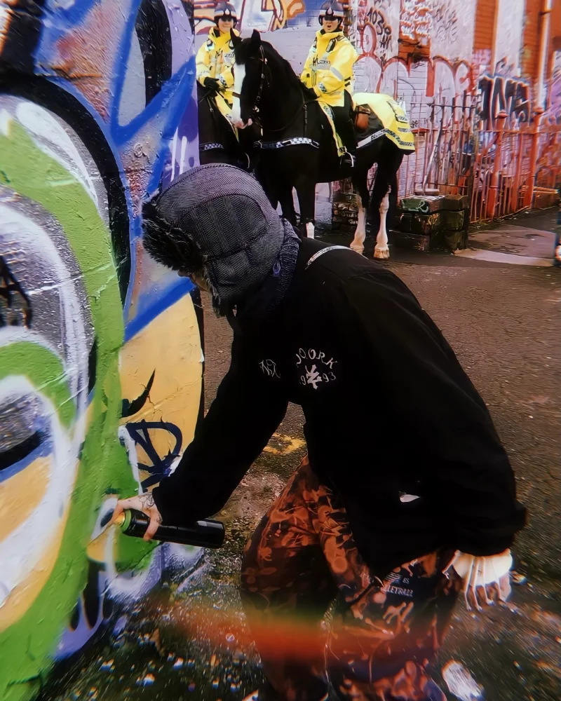 Kim spray painting a wall covered in graffiti with two police women stood behind her on horseback and one of them looks to be shouting at her. Kim is wearing streetwear with her hand in a complex plastic cast.