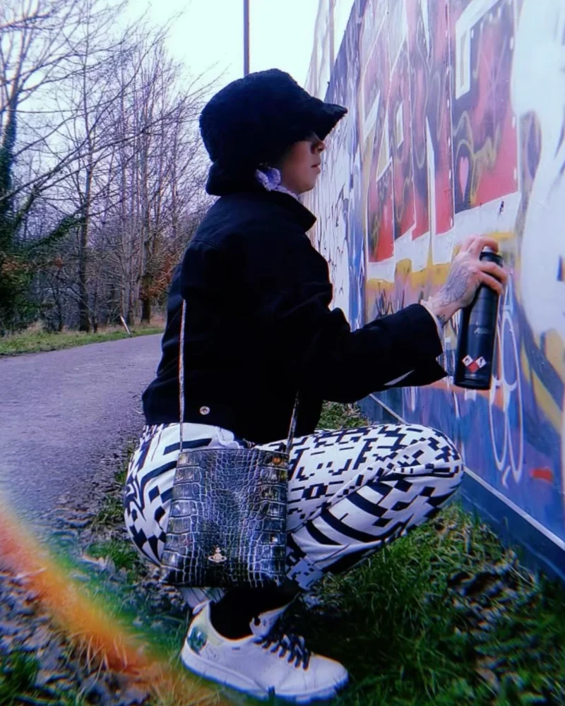 Kim wearing her own designs - a pair of black and white print tracksuit bottoms which look like a QR code - spray painting a wall which is covered in graffiti.