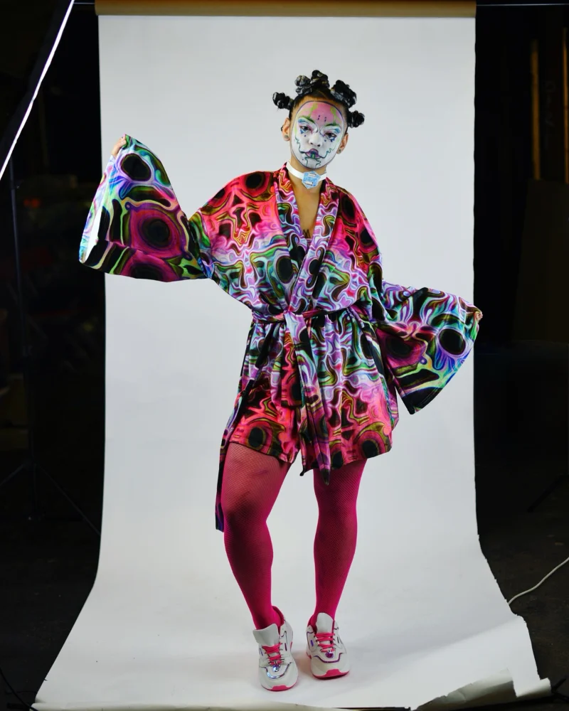 An alternative model with white futuristic geisha face-paint wears a Kimono featuring Kim's designs. Her hair is tied into lots of plaited buns. She wears pink tights and trainers. Standing on a white photographic backdrop.